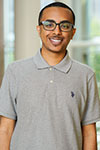Portrait of Naod Habtemariam standing indoors in front of a window, wearing a gray shirt and glasses