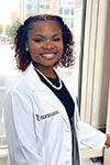 Portrait of Amiya Jenkins, standing indoors in front of a window, wearing a white coat and a strand of pearls